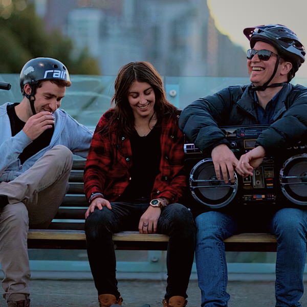 Friends sitting on a bench with the one on the right carrying a large radio speaker on his lap