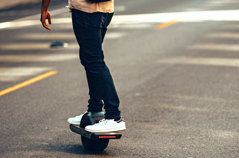 Cropped image of a man wearing a jeans and a white shoes riding a Onewheel Electric Skateboard across the road