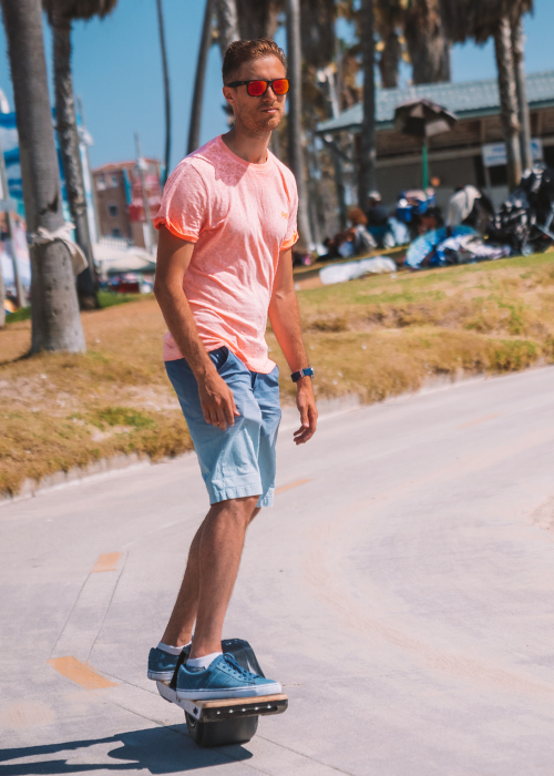 Man wearing a pink shirt and a sunglass rides a Onewheel Electric Skateboard on a curve road