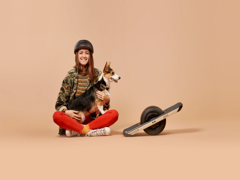 Woman holding a dog while sitting on the floor beside a Onewheel Pint electric skateboard