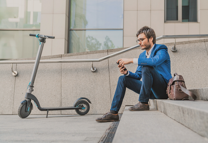 Young businessman sitting on stairs while using a phone near an Electric Scooter - My Electric Scooter