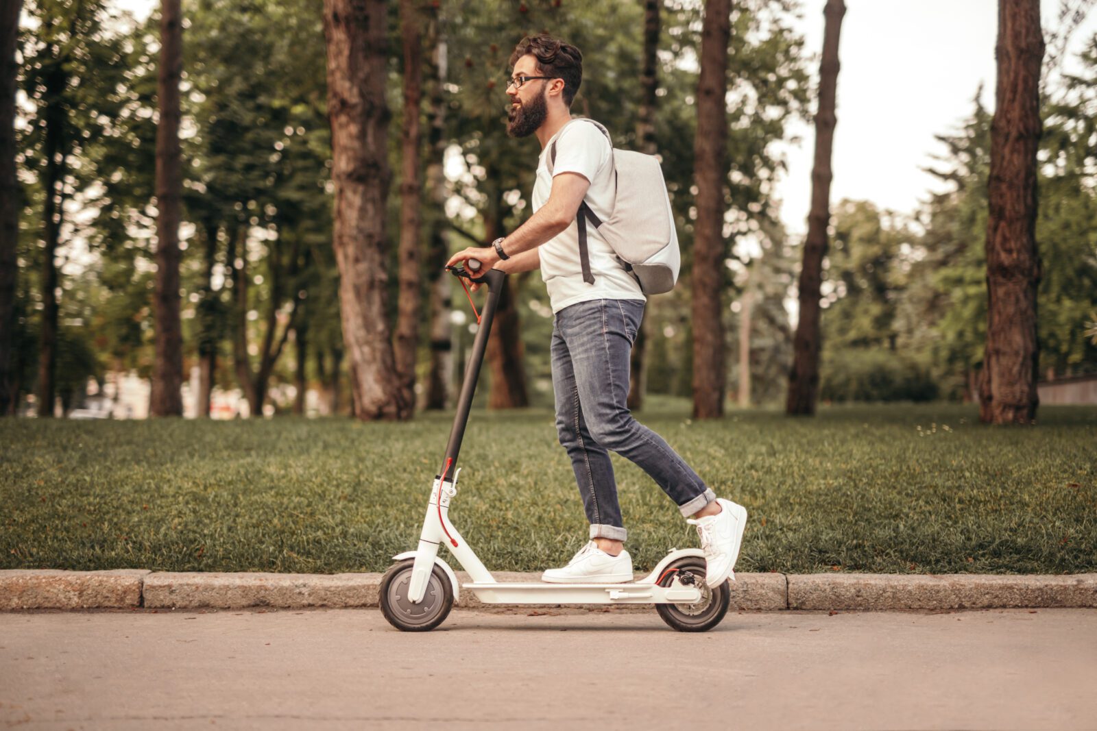 Side view of bearded man in casual clothes with backpack riding electric scooter in park - My Electric Scooter