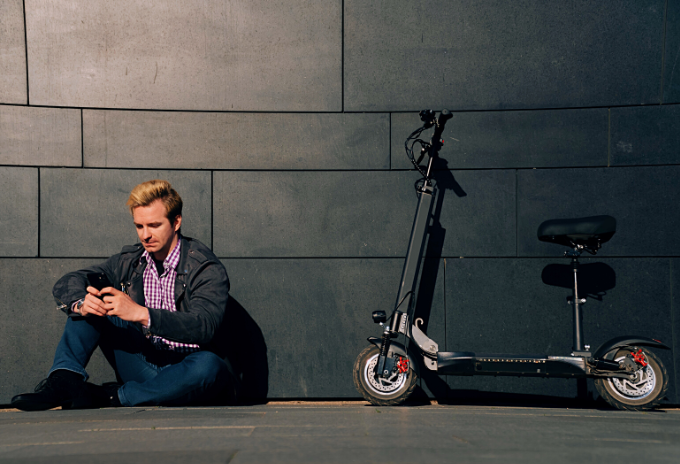 Man sitting on the floor using a smarthphone and an electric scooter with seat attached leaning against a wall