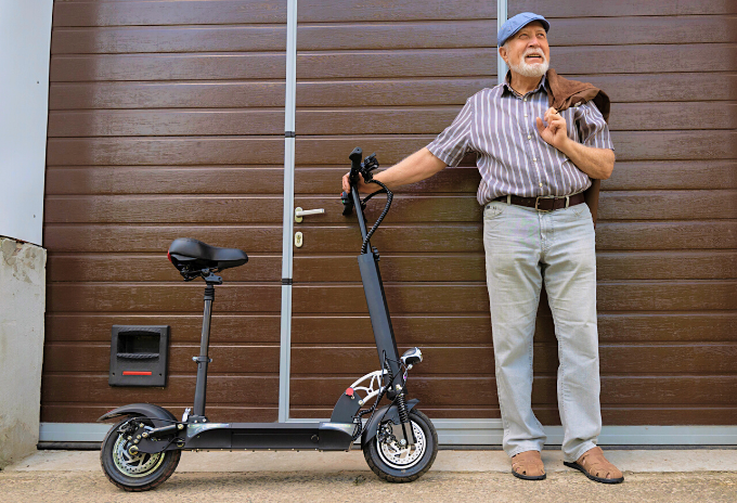 Old man holding to an electric scooter with seat in front of a gate