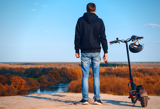 Man standing beside an electric off road scooter with helmet, river and trees can be seen on the background